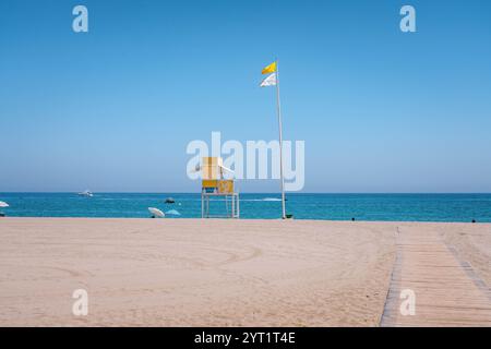 Gelber Rettungsschwimmerturm mit Fahnen an einem Sandstrand mit einigen Booten in der Ferne und blauem Himmel Stockfoto