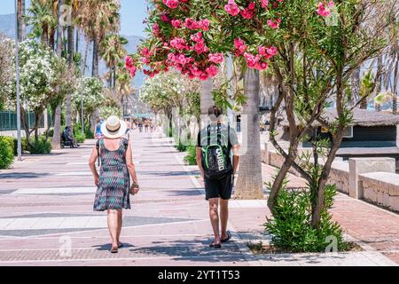 Touristen schlendern entlang einer gepflasterten Promenade, die von Palmen und leuchtenden rosa Oleanderblüten gesäumt ist, und genießen einen sonnigen Tag am Meer Stockfoto