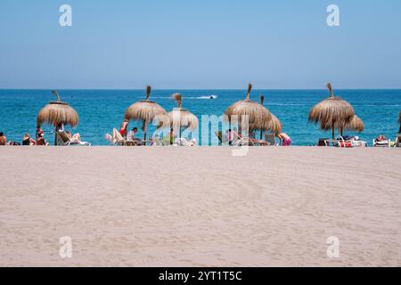 Die Leute genießen einen sonnigen Tag am Strand unter Strohschirmen, liegen auf Liegen nahe dem klaren blauen Meer mit einem Boot in der Ferne Stockfoto