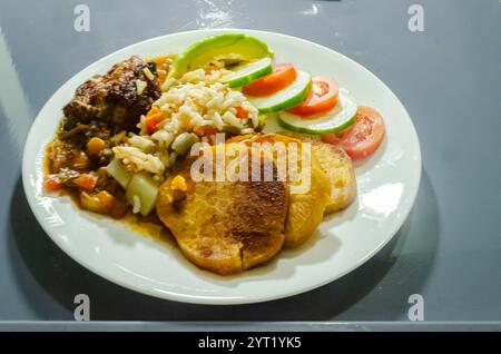 Tropisches jamaikanisches Gericht mit kandierter Süßkartoffel, Reis, Bohnen, Karotten, Huhn, Tomaten, Gurken und Avocado auf einem Teller auf einem Tablett serviert. Stockfoto