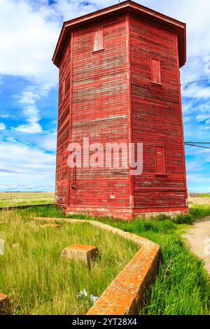 Ein roter Backsteinturm steht auf einem Feld mit einer grünen Grasfläche um ihn herum. Der Turm ist alt und hat ein verwittertes Aussehen. Der Himmel ist klar und blau, an Stockfoto