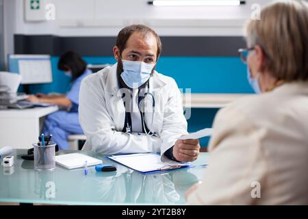 Maskierter Arzt mit Gesichtsmaske, der älteren Frau in der Arztpraxis verschreibungspflichtiges Papier gibt. Rentnerin-Patientin erhält schriftlichen Behandlungsplan von jungen Hausärzten. Stockfoto