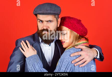 Herbst-Winter-Mode. Hübscher bärtiger Mann im Anzug und Mütze, der die schöne Frau in einem Wollpullover, einer roten Baskenmütze und einem farbenfrohen Schal umarmt. Romantische junge Co Stockfoto