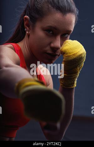 Entschlossene Boxerin, die Handumschläge trägt und während eines intensiven Trainings in einem schwach beleuchteten Fitnessstudio einen kräftigen Schlag auf den Zuschauer wirft, Dämon Stockfoto