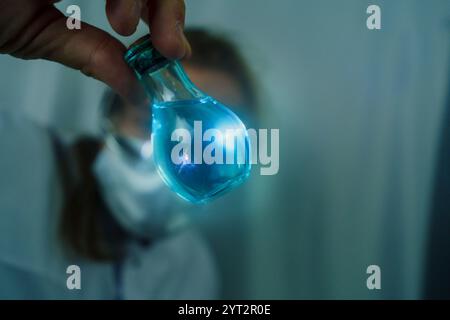 Ein Wissenschaftler, der eine Schutzbrille trägt, hält einen Rundkolben mit leuchtender blauer Flüssigkeit in einem Labor. Chemie-Experiment, Forschung und Wissenschaft Stockfoto