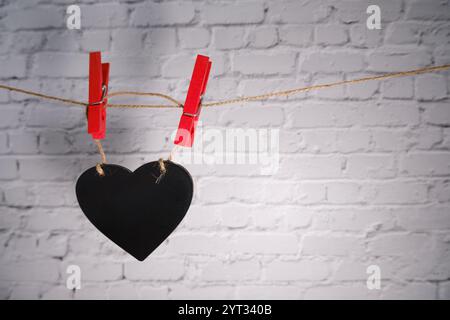 Schwarzes Herz, das an einer Schnur hängt, mit roten Wäschespins, Backsteinmauerhintergrund. Stockfoto