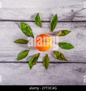 Ein quadratisches Foto mit einer Mandarine und grünen Blättern, die in Form einer Blume auf einem grauen Holztisch angeordnet sind. Stockfoto