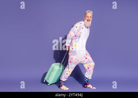 Fröhlicher reifer Mann im Einhorn-Pyjama mit Koffer auf leuchtendem lila Hintergrund Stockfoto