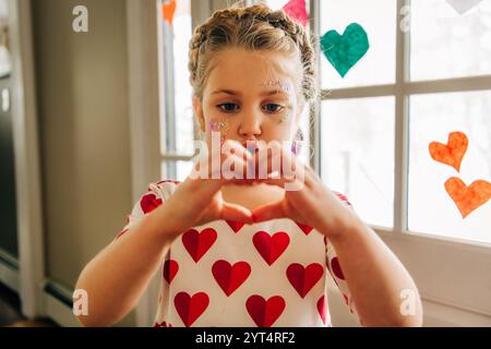 Mädchen im Kleid mit Herzmuster bildet Herzform mit Händen in der Nähe des Fensters Stockfoto