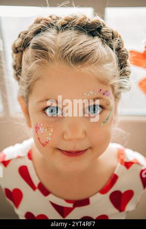 Mädchen mit geflochtenen Haaren und Glitzer-Herz-Make-up lächelt sanft Stockfoto