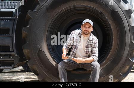 Männlicher Mechaniker mit digitalem Tablet, der in einem großen Rad der Bau- oder Landmaschine sitzt. Stockfoto