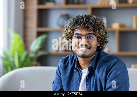 Ein junger Mann mit lockigen Haaren und Brille sitzt auf einem Sofa und lächelt warm. Er trägt ein lässiges blaues Hemd, was eine freundliche und zugängliche Atmosphäre in einem modernen Wohnbereich schafft. Stockfoto