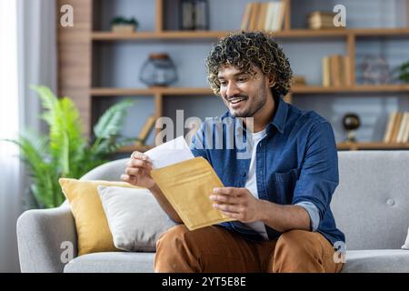Ein fröhlicher Mann wird gesehen, wie er einen Umschlag öffnet, der bequem auf einem Sofa sitzt. Sein Ausdruck spiegelt Aufregung und Eifer wider. Das Ambiente ist ein gemütliches Wohnzimmer, das Wärme ausstrahlt. Stockfoto