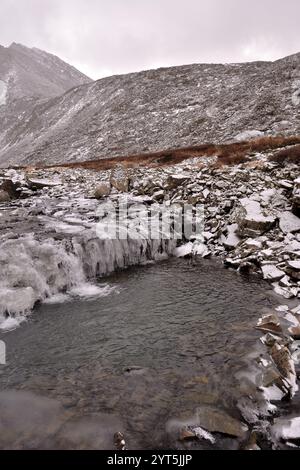 Ein teilweise gefrorener Bach eines flachen Flusses fließt in ein kleines Rückwasser, das von hohen Bergen umgeben ist, im ersten Schnee an einem bewölkten Herbsttag. Naryn Stockfoto