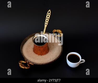 Ein Tablett mit Sand, in dem ein Kupfertürke mit schwarzem Kaffee auf schwarzem Hintergrund steht, daneben eine Tasse mit einem heißen Getränk. Nahaufnahme. Stockfoto