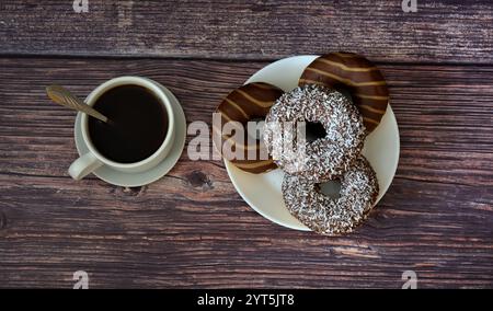 Ein Teller mit mehreren Donuts in Schokolade und Karamellglasur mit Streuseln und einer Tasse schwarzen Kaffee mit Untertasse auf einem dunklen Holztisch. Draufsicht, fl Stockfoto