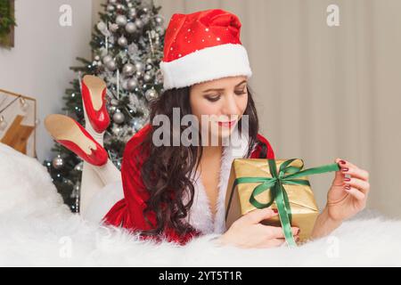 Weihnachtsgeschenk - Frau Eröffnungsgeschenk überrascht und glücklich, Junge schöne lächelnde Frau in Santa Hut. Lustiges, süßes Foto von Frau auf dem Bett im Morni Stockfoto