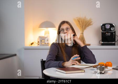 Ein traumhaftes Mädchen schreibt einen Brief an den Weihnachtsmann, um sich auf Weihnachten vorzubereiten. Sie sitzt an einem Tisch mit Notizbuch und Stift in der Küche. Stockfoto