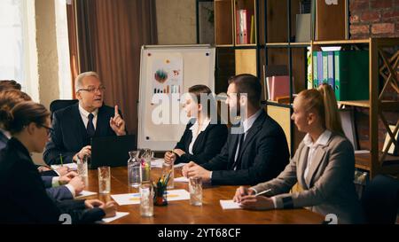 Ein leitender Geschäftsmann präsentiert Ideen während eines Unternehmenstreffens mit jungen Kollegen und betont dabei die wichtigsten Punkte, um das Team mit unterstützenden Datendiagrammen zu unterstützen. Stockfoto