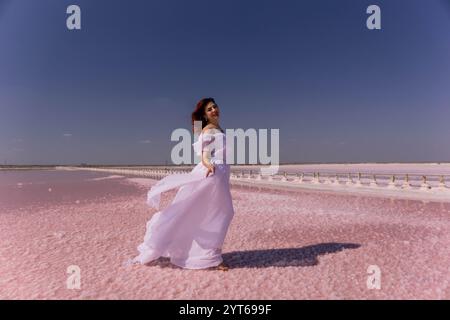 Woman White Dress Pink Salt Lake – Eine Frau in einem weißen Kleid steht auf einem rosa Salzsee. Die Frau schaut nach rechts und der Wind weht sie Stockfoto
