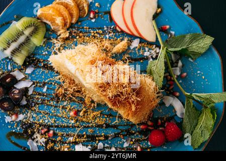 Elegantes Dessert mit Kunafa und frischen Obstscheiben auf blauem Teller. Eine kreative Präsentation von Süßigkeiten aus dem Nahen Osten mit lebhaften Beilagen. Stockfoto