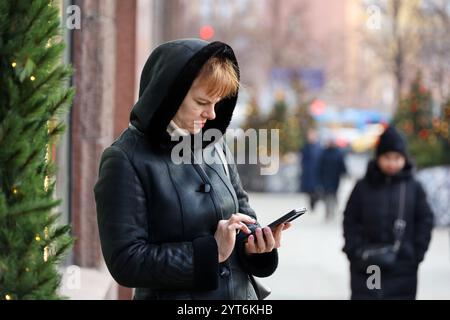 Frau mit warmem Mantel mit Smartphone auf der Stadtstraße auf dem Hintergrund von Neujahrsbäumen, mobile Kommunikation im Winter Stockfoto