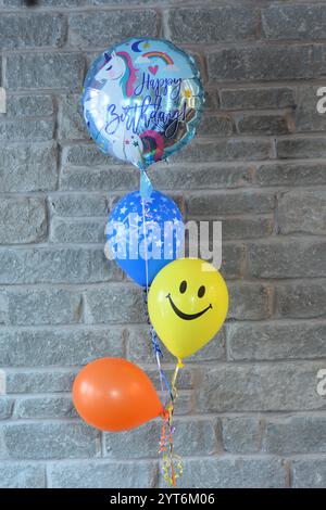 Vier bunte Helium Happy Birthday Ballons schweben auf einer Schnur vor einer grauen Ziegelwand in einem Haus, bereit für eine Geburtstagsfeier. Stockfoto