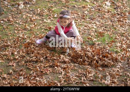 Ein junges Mädchen mit rosa Weste und Regenbogenstiefeln sammelt an einem sonnigen Nachmittag in einem Park Herbstlaub Stockfoto