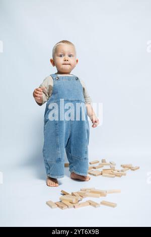 Niedlicher Junge in Denim Latzhose auf weißem Hintergrund. Kind, das mit Holzspielzeug spielt. Lächelndes Kleinkind in toller Stimmung Stockfoto