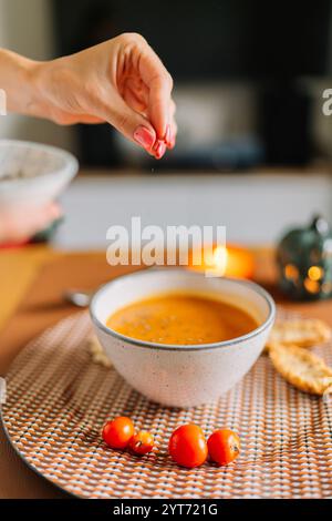 Von Hand mit Gewürz über eine Schüssel Kürbissuppe mit Tomaten bestreuen. Nahaufnahme der Küchenchefin, die gekochte Suppe in der rustikalen Küche salzt. Stockfoto