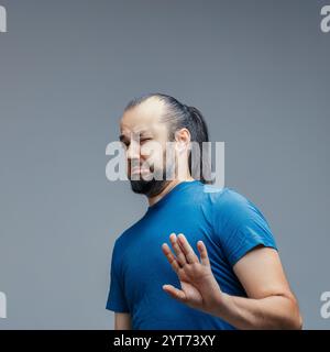 Mann mit langen dunklen Haaren im Pferdeschwanz, mit blauem T-Shirt, mit angeekeltem Gesicht und abstoßender Hand vor grauem Hintergrund Stockfoto