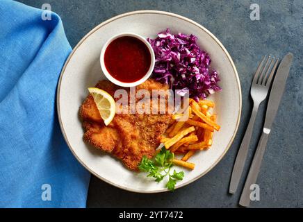 Schnitzel mit Kartoffelfrites, Rotkohlsalat und Sauce auf weißem Teller auf dunklem Steinhintergrund. Draufsicht, flach Stockfoto