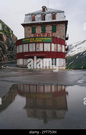 Blick auf das Hotel Belvedere am Furka Pass in der Schweiz. Das berühmte Steingebäude spiegelt sich in einer Pfütze am Straßenrand wider. Stockfoto