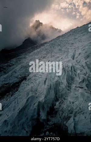 Die ersten Sonnenstrahlen des Tages brechen durch den Nebel und erhellen den Gipfel der Aiguille du Midi bei Chamonix Mont-Blanc. Im Vordergrund steht der Bossons-Gletscher mit seinen Türmen und Eisspalten. Die Atmosphäre ist mystisch und düster. Ein Vogel fliegt am Bildrand. Stockfoto