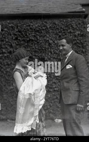 1930er Jahre, historisch, Taufe, Paar, eine junge Dame, die ihr Baby in ein Seidenkleid gewickelt hält. Stockfoto