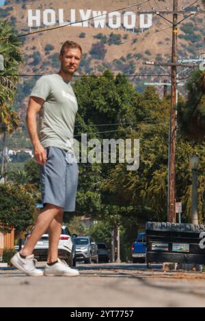 Ein Mann läuft eine von Bäumen gesäumte Straße in Los Angeles, Kalifornien, entlang, mit dem berühmten Hollywood-Schild im Hintergrund, umgeben von geparkten Autos. Stockfoto