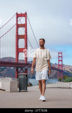 Ein Mann in einem leichten T-Shirt und einer kurzen Hose spaziert über einen Weg mit den rot-orangen Türmen der Golden Gate Bridge und Kabeln, die vor einem teilweise bewölkten s zu sehen sind Stockfoto