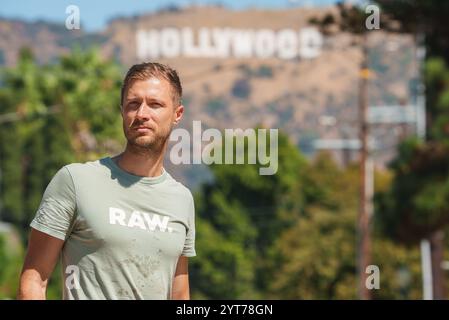 Ein Mann steht draußen und trägt ein hellgrünes T-Shirt mit RAW-Aufdruck. Das berühmte Hollywood-Schild ist sichtbar, umgeben von Grün und Palmen Stockfoto