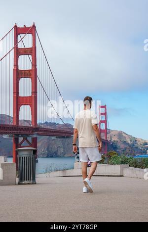 Eine Person läuft in Richtung der Golden Gate Bridge in San Francisco, Kalifornien. Die rot-orangen Türme heben sich von einem blauen Himmel mit verstreuten Wolken ab. Stockfoto