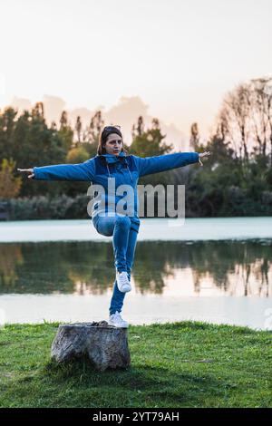 Junge Frau, die beim Sonnenuntergang auf einem Baumstumpf am See Yoga praktiziert und die Ruhe der Natur genießt Stockfoto