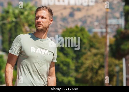 Ein Mann steht draußen und trägt ein hellgrünes T-Shirt mit RAW-Aufdruck. Das berühmte Hollywood-Schild ist sichtbar, umgeben von Grün und Palmen Stockfoto