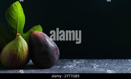 Minimalistische Komposition mit zwei Reifen Feigen neben einem leuchtend grünen Feigenblatt, vor dunklem Hintergrund mit einer strukturierten Oberfläche für eine Erde Stockfoto