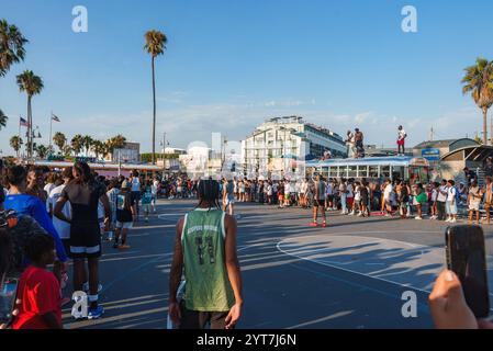 Eine lebhafte Szene am Venice Beach, Los Angeles, mit einem Basketballspiel auf einem Platz im Freien. Hohe Palmen und die belebte Promenade sind zu sehen Stockfoto