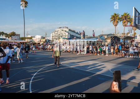 Eine lebhafte Szene am Venice Beach, Los Angeles, mit einer Menge um einen Basketballplatz, Palmen und einzigartiger Architektur unter einem klaren blauen Himmel. Stockfoto