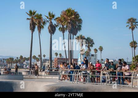 Am Venice Beach, Los Angeles, versammeln sich die Leute an einem Geländer und beobachten die Eisläufer in einem Park. Hohe Palmen und ein klarer blauer Himmel unterstreichen den sonnigen Tag. Stockfoto