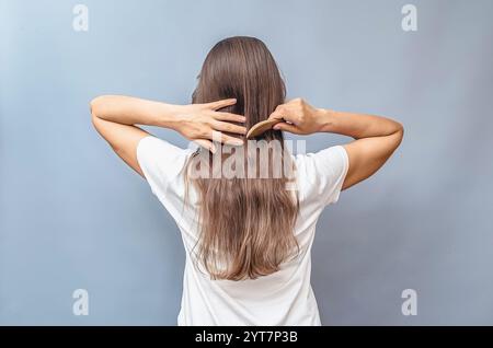 Kämmen von natürlichem langem Haar. Rückansicht einer Frau im weißen T-Shirt. Grauer Hintergrund, isoliert. Holzkamm Stockfoto