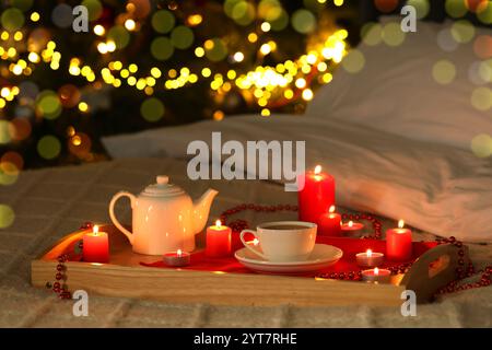 Aromatischer Tee in der Tasse, Teekanne und brennende Kerzen auf dem Bett gegen verschwommenes Weihnachtslicht, Bokeh-Effekt Stockfoto