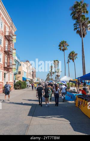 Die Leute laufen entlang der belebten Promenade Venice Beach, gesäumt von Palmen und farbenfrohen Verkaufsständen. Ein rotes Backsteingebäude mit Feuerflüchten ist Vis Stockfoto