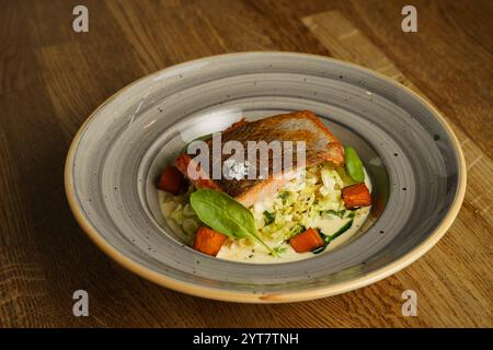 Ein perfekt gegrilltes Lachsfilet, serviert mit geröstetem Gemüse und einer cremigen Sauce auf einem dekorativen Teller. Das Gericht ist mit Spinatblättern garniert, Stockfoto