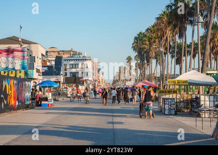 Die Menschen spazieren und radeln entlang des lebendigen Venice Beach Boardwalk in Los Angeles. Palmen säumen den Weg, mit farbenfrohen Graffiti-Kunst an den Wänden in der Nähe. Stockfoto